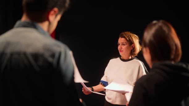 A female theater play director is giving instructions to a group of performers on stage. A group of theater stage performers are checking texts and rehearsing.